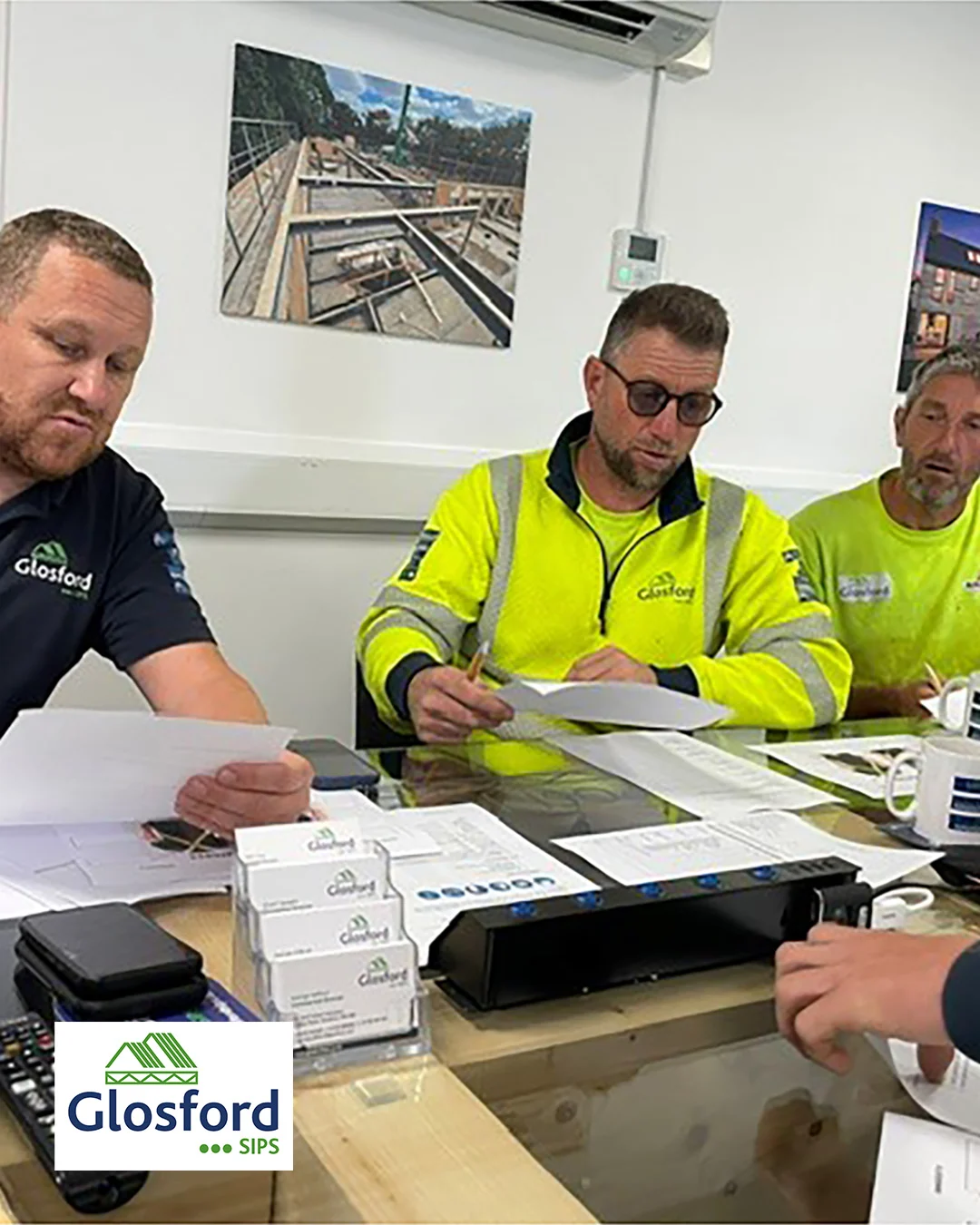 Three construction workers in high-visibility Glosford shirts reviewing paperwork around a table during a site meeting.