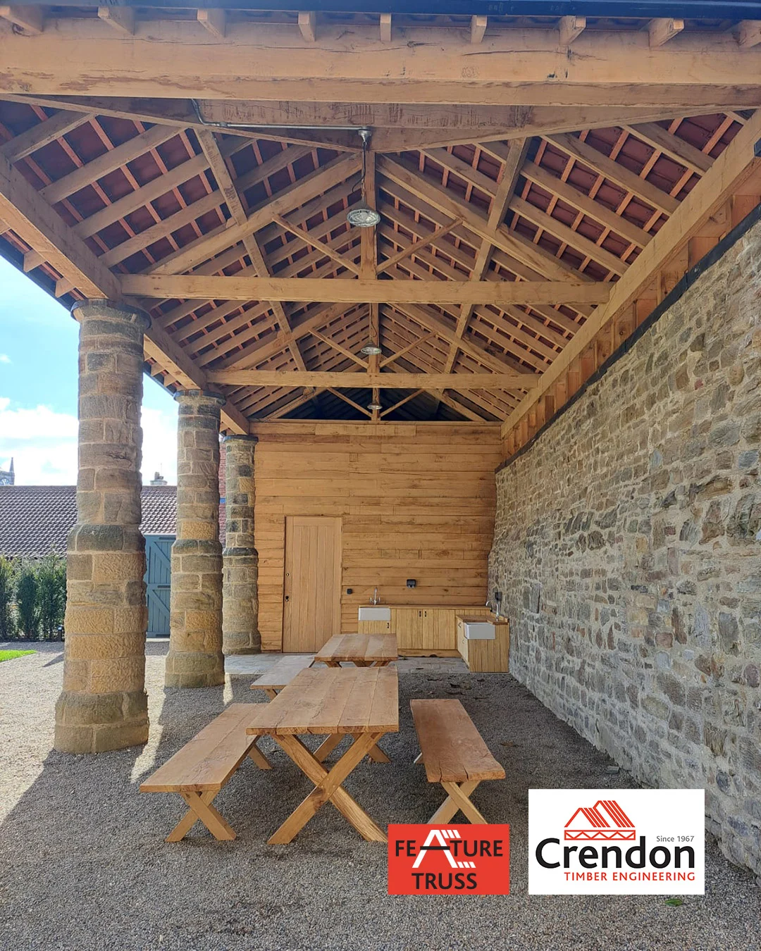 Open timber pavilion with exposed timber trusses, stone columns and wall, picnic benches and service counters; Crendon logo in bottom right