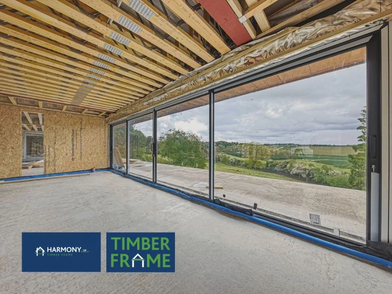 Unfinished timber-frame room with exposed joists and floor-to-ceiling glass overlooking rolling countryside; Harmony logo.