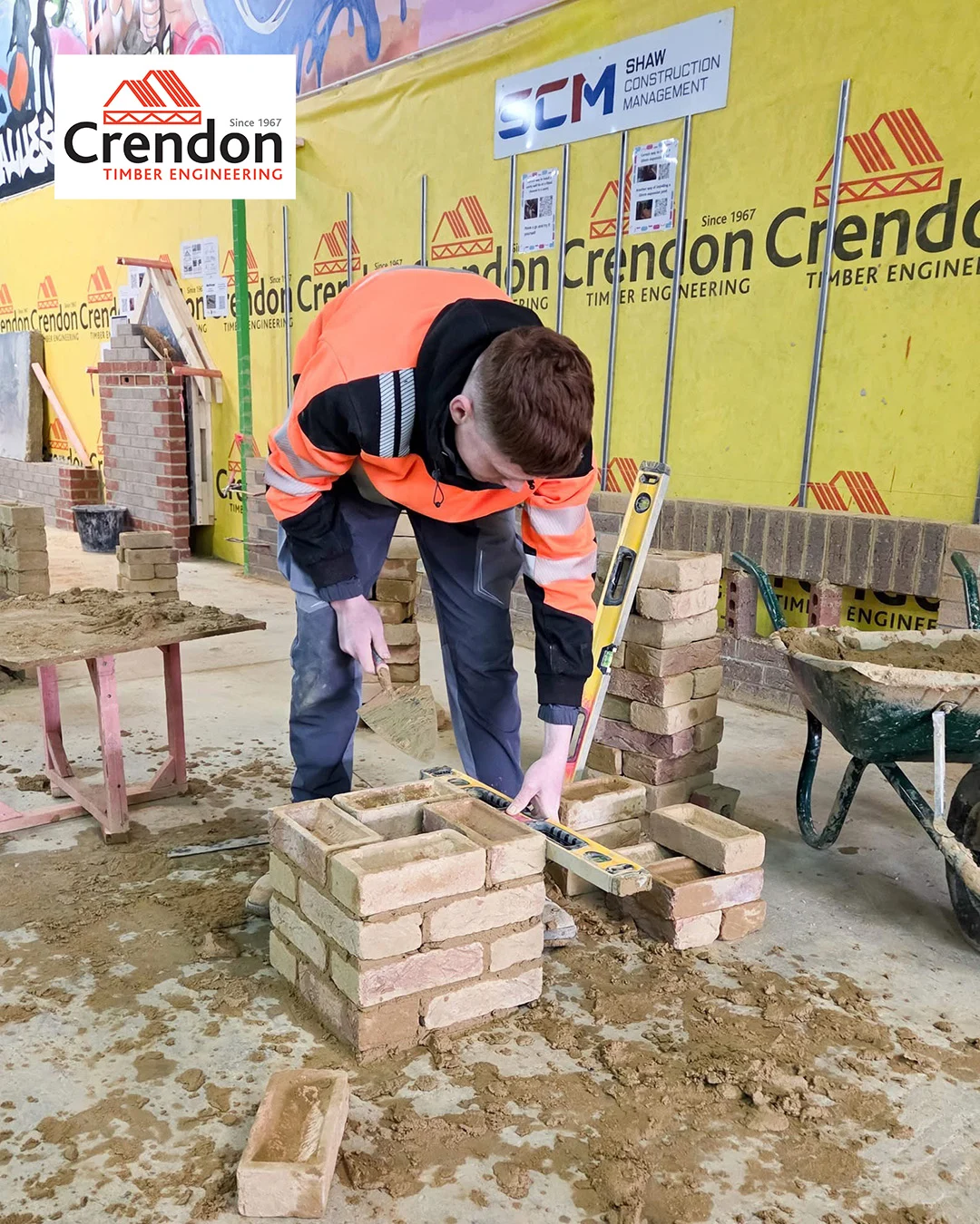 Worker in orange hi-vis jacket laying bricks with trowel and spirit level in indoor masonry workshop with Crendon signage.